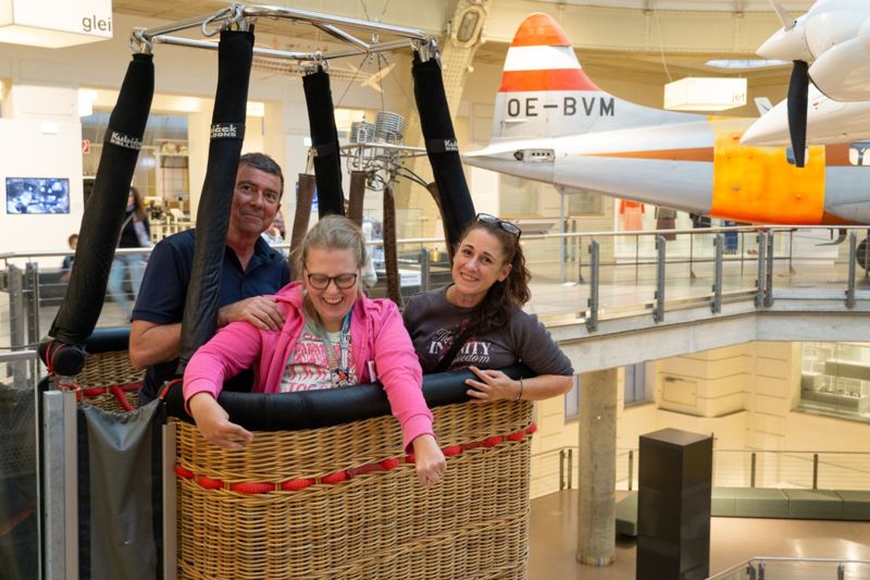 Drei Personen stehen lachend in einem Ballonkorb im Museum.