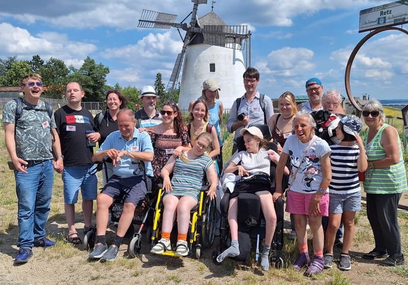Eine große Gruppe, viele im Rollstuhl, vor einer weißen Windmühle.