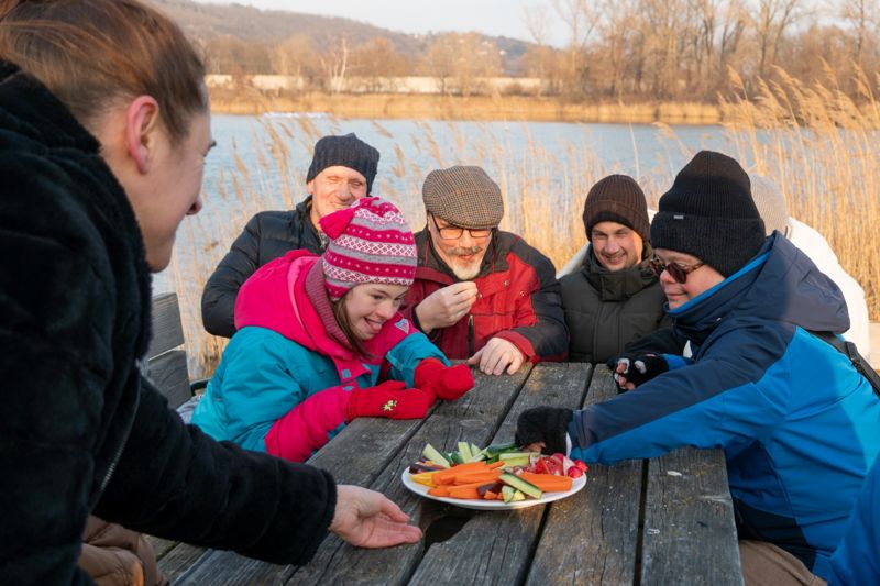 Eine Gruppe in Winterkleidung macht Jause an einem Holztisch am See.
